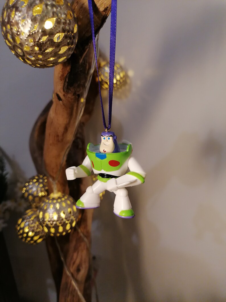 May include: A white, green and purple plastic ornament of Buzz Lightyear from the Toy Story franchise. The ornament is hanging from a purple ribbon.