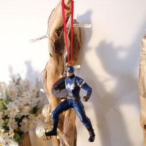 May include: A red ribbon hangs from a piece of driftwood, with a Captain America action figure ornament attached. The figure is in a flying pose, holding his shield.