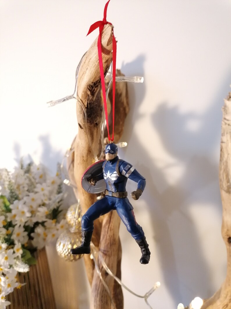 May include: A red ribbon hangs from a piece of driftwood, with a Captain America action figure ornament attached. The figure is in a flying pose, holding his shield.