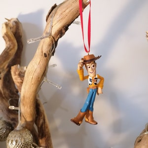 May include: A red ribbon hangs from a branch with a plastic figurine of Woody from Toy Story. Woody is wearing his signature cowboy hat, vest, and jeans. He is holding his hat in his left hand and has a friendly expression on his face.