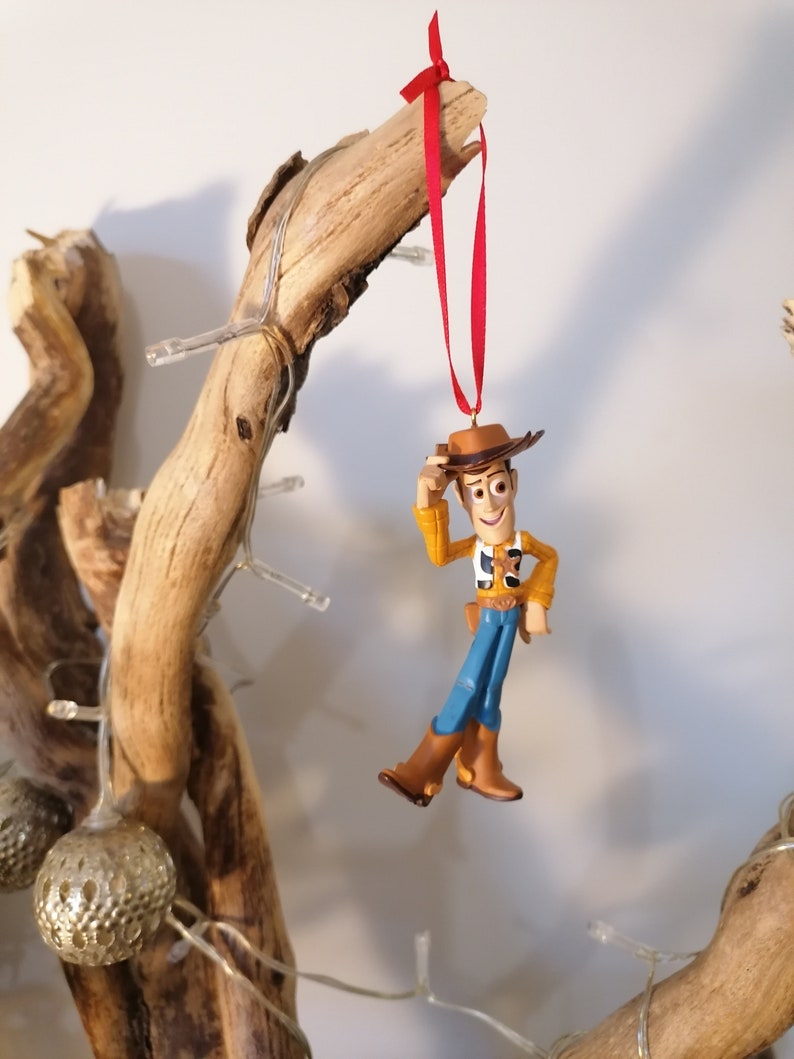 May include: A red ribbon hangs from a branch with a plastic figurine of Woody from Toy Story. Woody is wearing his signature cowboy hat, vest, and jeans. He is holding his hat in his left hand and has a friendly expression on his face.