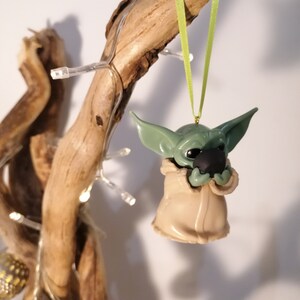 May include: A green and brown Yoda ornament hanging from a branch with a green ribbon. The ornament is holding a small, round, black object.