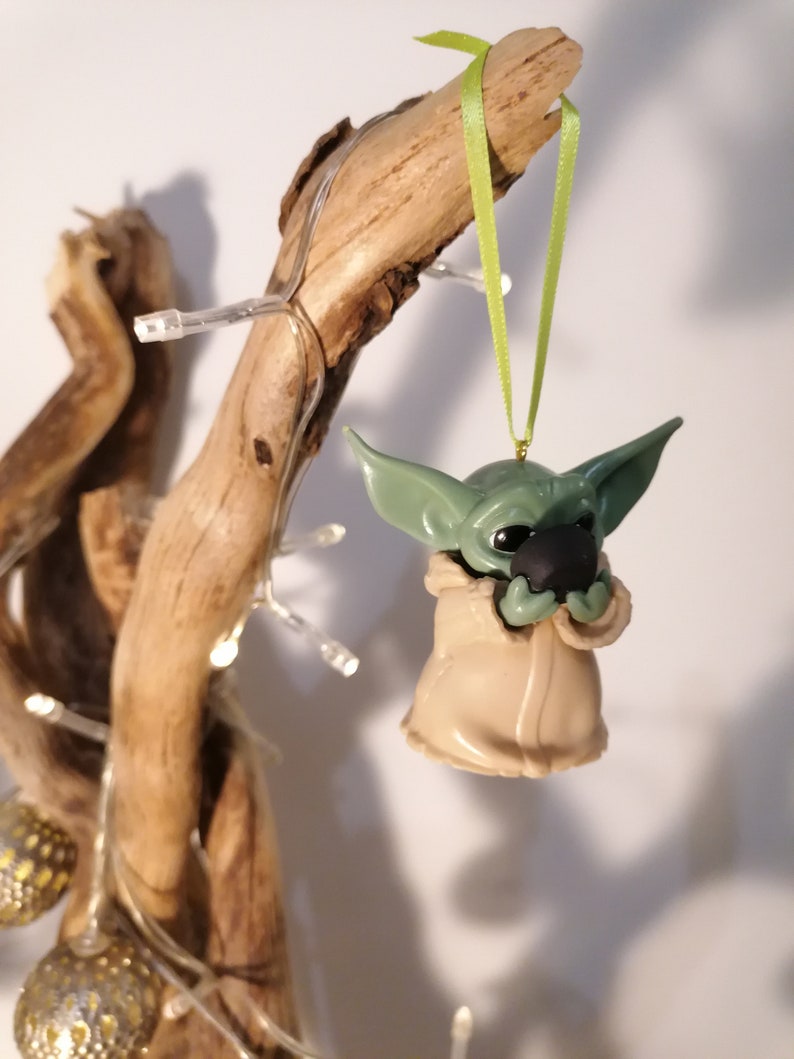 May include: A green and brown Yoda ornament hanging from a branch with a green ribbon. The ornament is holding a small, round, black object.