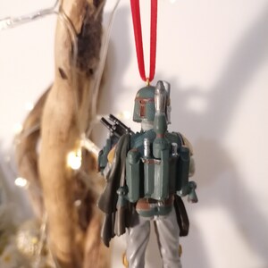 May include: A green and gray Star Wars Boba Fett Christmas ornament with a red ribbon. The ornament is a detailed figurine of Boba Fett with his signature jetpack and blaster.