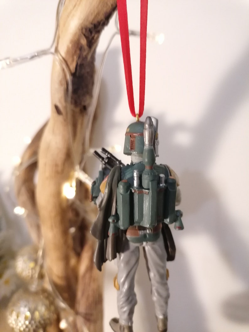May include: A green and gray Star Wars Boba Fett Christmas ornament with a red ribbon. The ornament is a detailed figurine of Boba Fett with his signature jetpack and blaster.