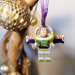 May include: A purple and white Christmas bauble featuring a plastic figurine of Buzz Lightyear from the Toy Story franchise. The bauble is hanging from a purple ribbon.