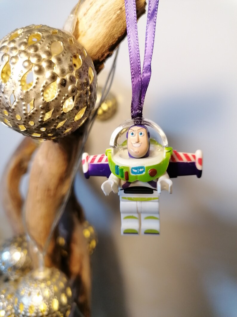May include: A purple and white Christmas bauble featuring a plastic figurine of Buzz Lightyear from the Toy Story franchise. The bauble is hanging from a purple ribbon.