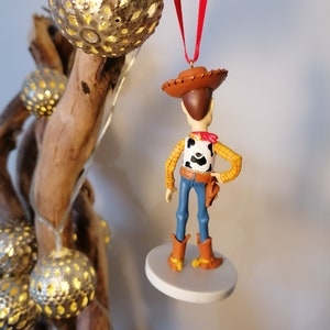 Op de afbeelding: Een rode draad hangt aan een witte ronde basis met een klein plastic beeldje van Woody uit Toy Story. Woody draagt zijn kenmerkende cowboyhoed, vest en jeans. Hij staat met zijn rug naar de camera.