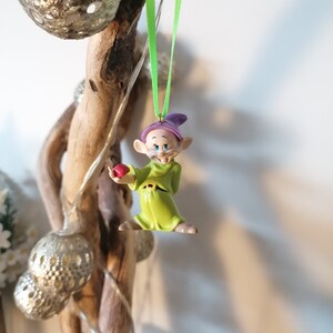 May include: A green and purple plastic figurine of a dwarf holding a red apple. The dwarf is wearing a green tunic and a purple hat. The figurine is hanging from a green ribbon.