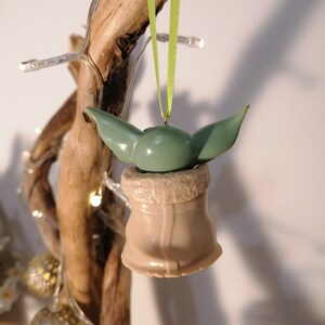 May include: A green and brown Christmas ornament shaped like a character from the Star Wars franchise. The ornament is hanging from a green ribbon on a branch.