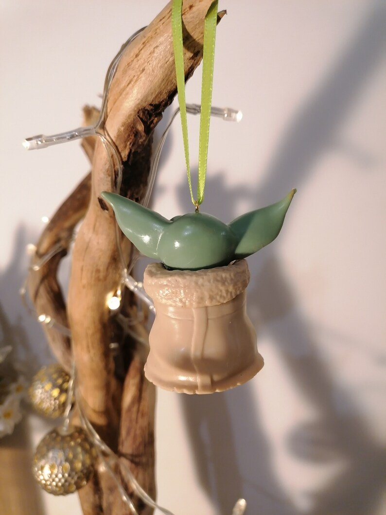 May include: A green and brown Christmas ornament shaped like a character from the Star Wars franchise. The ornament is hanging from a green ribbon on a branch.