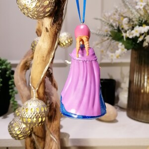 Puede incluir: Un adorno navide&ntilde;o morado y azul con una ni&ntilde;a con el pelo largo trenzado que lleva una capa morada. El adorno est&aacute; colgado de una cinta azul.