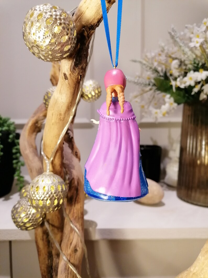 Puede incluir: Un adorno navide&ntilde;o morado y azul con una ni&ntilde;a con el pelo largo trenzado que lleva una capa morada. El adorno est&aacute; colgado de una cinta azul.