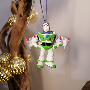 Puede incluir: Un adorno de juguete de plástico blanco y verde de Buzz Lightyear de la franquicia Toy Story. El adorno está colgado de una cinta morada y tiene un traje de ranger espacial blanco y verde con un chaleco morado. Buzz Lightyear tiene un casco blanco con una visera transparente y un logotipo de ranger espacial verde y blanco en el pecho.