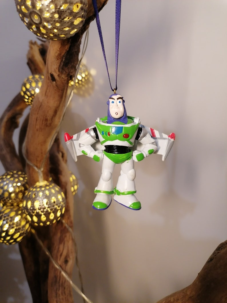 Pu&ograve; includere: Un ornamento di giocattolo in plastica bianca e verde di Buzz Lightyear della serie Toy Story. L'ornamento &egrave; appeso a un nastro viola e ha una tuta da ranger spaziale bianca e verde con un gilet viola. Buzz Lightyear ha un casco bianco con una visiera trasparente e un logo da ranger spaziale verde e bianco sul petto.