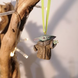 Pu&ograve; includere: Una statuetta verde e marrone di un personaggio del franchise di Star Wars, che indossa un cappotto marrone, appesa a un nastro verde. La statuetta &egrave; un ornamento natalizio.