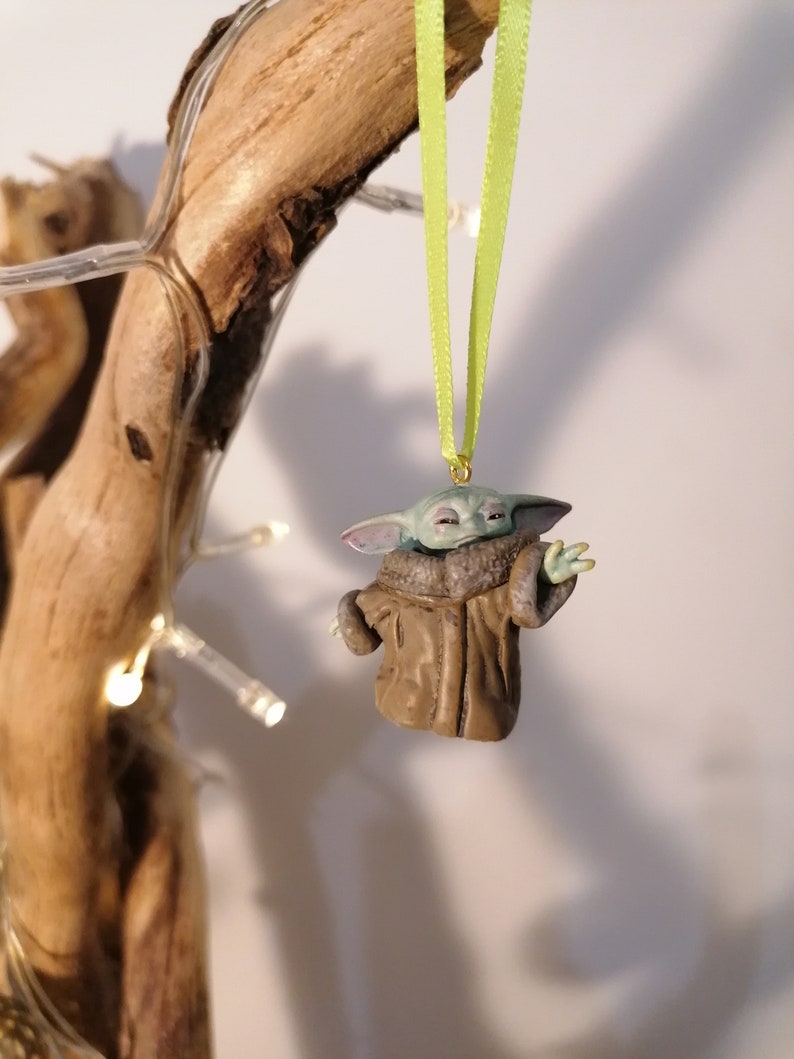 Pu&ograve; includere: Una statuetta verde e marrone di un personaggio del franchise di Star Wars, che indossa un cappotto marrone, appesa a un nastro verde. La statuetta &egrave; un ornamento natalizio.