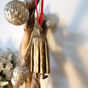 May include: A gold-colored ornament of a person wearing a cape, hanging from a red ribbon on a branch.