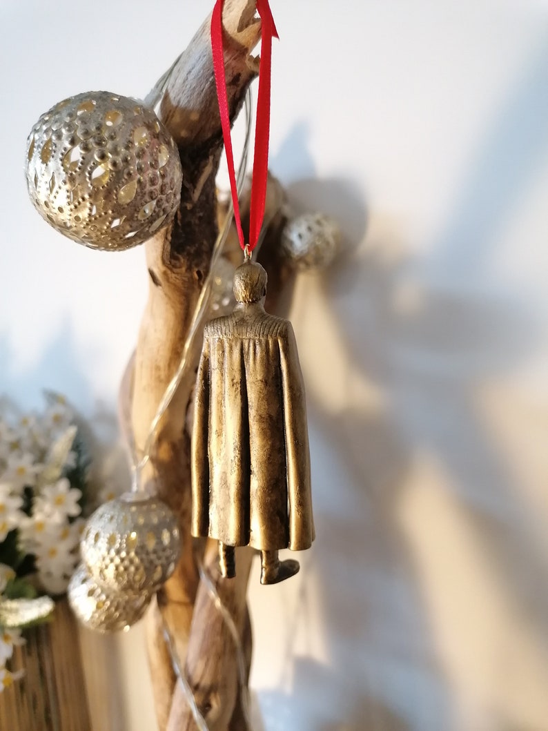 May include: A gold-colored ornament of a person wearing a cape, hanging from a red ribbon on a branch.