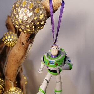 May include: A green and white plastic toy figurine of Buzz Lightyear from the Toy Story movie franchise hanging from a purple ribbon. The figurine is wearing a space ranger uniform with a green vest and a white helmet.