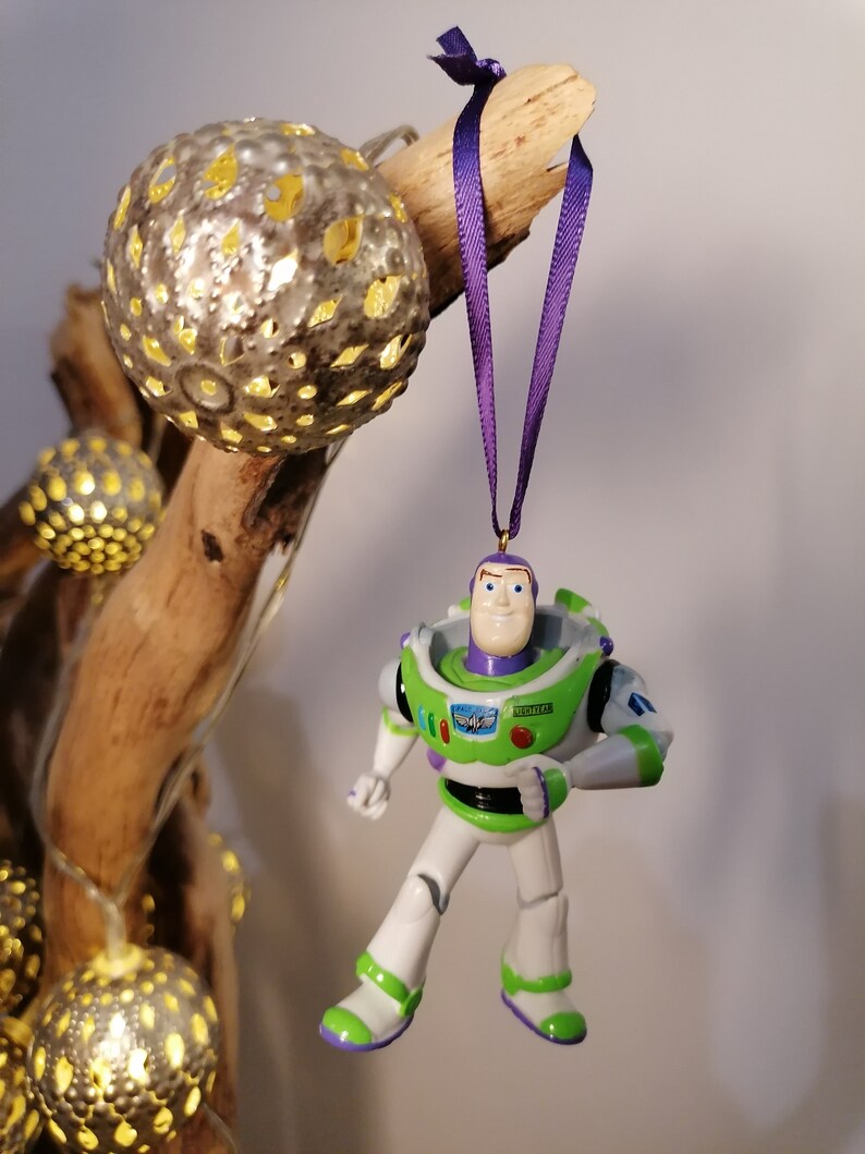 May include: A green and white plastic toy figurine of Buzz Lightyear from the Toy Story movie franchise hanging from a purple ribbon. The figurine is wearing a space ranger uniform with a green vest and a white helmet.