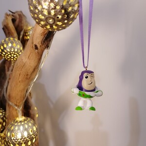 May include: Purple and white plastic toy ornament of Buzz Lightyear from the Toy Story franchise, hanging from a purple ribbon on a branch with string lights.