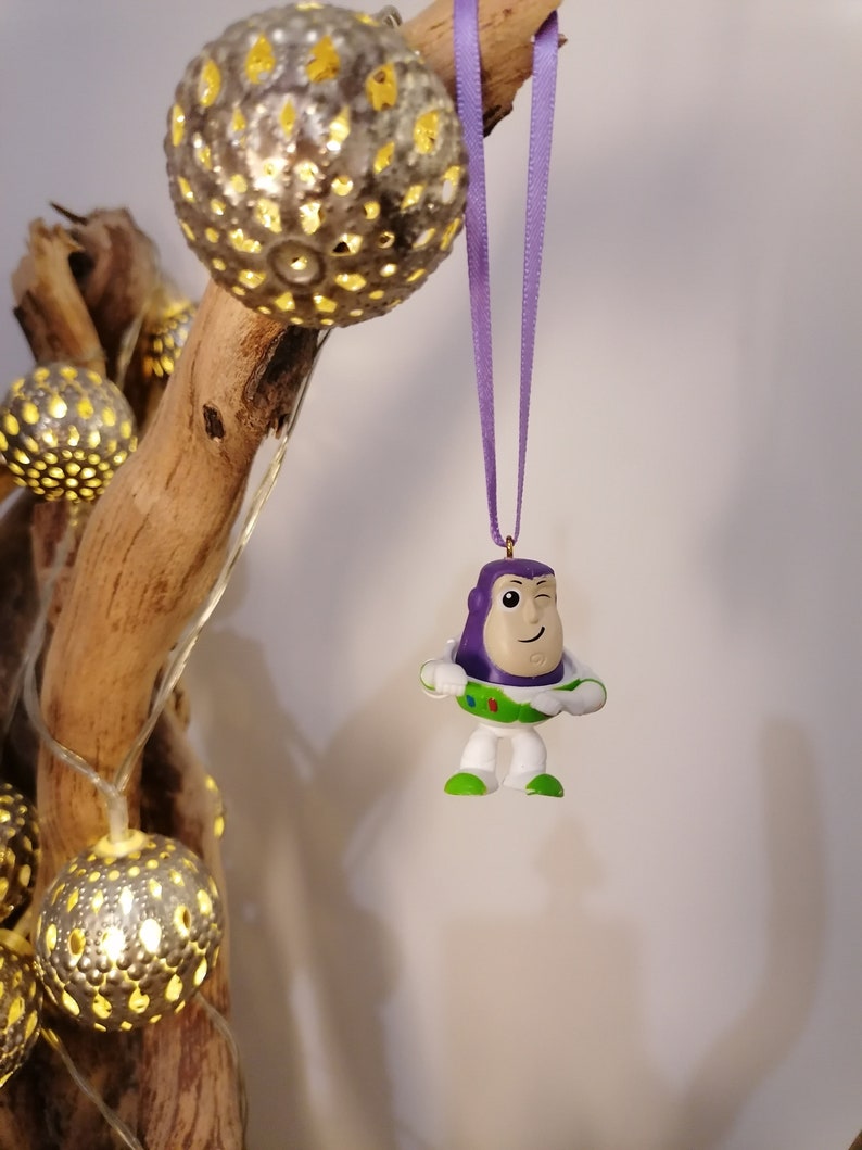 May include: Purple and white plastic toy ornament of Buzz Lightyear from the Toy Story franchise, hanging from a purple ribbon on a branch with string lights.