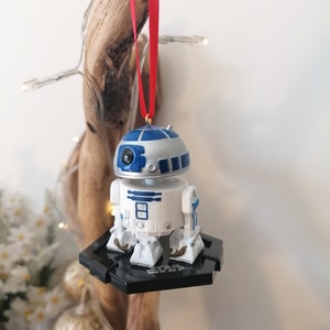 May include: A white and blue Star Wars R2-D2 Christmas ornament with a red ribbon. The ornament is hanging from a piece of driftwood.