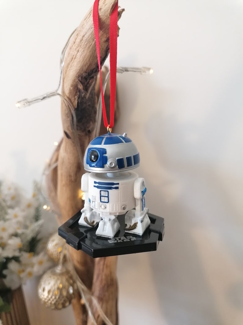 May include: A white and blue Star Wars R2-D2 Christmas ornament with a red ribbon. The ornament is hanging from a piece of driftwood.
