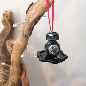 K&ouml;nnte beinhalten: Schwarzer und silberner Star Wars BB-8-Anh&auml;nger mit einem roten Band, das an einem Ast h&auml;ngt. Der Anh&auml;nger befindet sich auf einem schwarzen Sockel mit der Aufschrift "Star Wars".