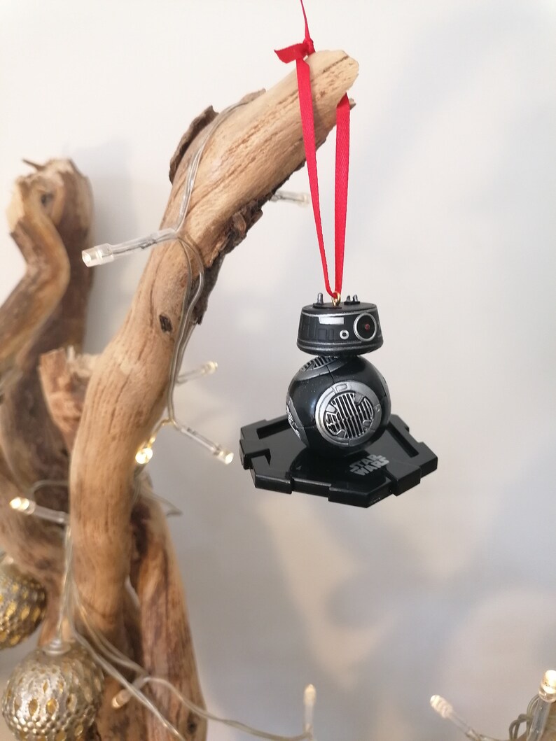 K&ouml;nnte beinhalten: Schwarzer und silberner Star Wars BB-8-Anh&auml;nger mit einem roten Band, das an einem Ast h&auml;ngt. Der Anh&auml;nger befindet sich auf einem schwarzen Sockel mit der Aufschrift "Star Wars".