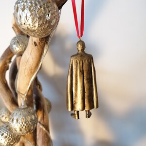 May include: A gold-colored ornament of a person wearing a cape, hanging from a red ribbon.