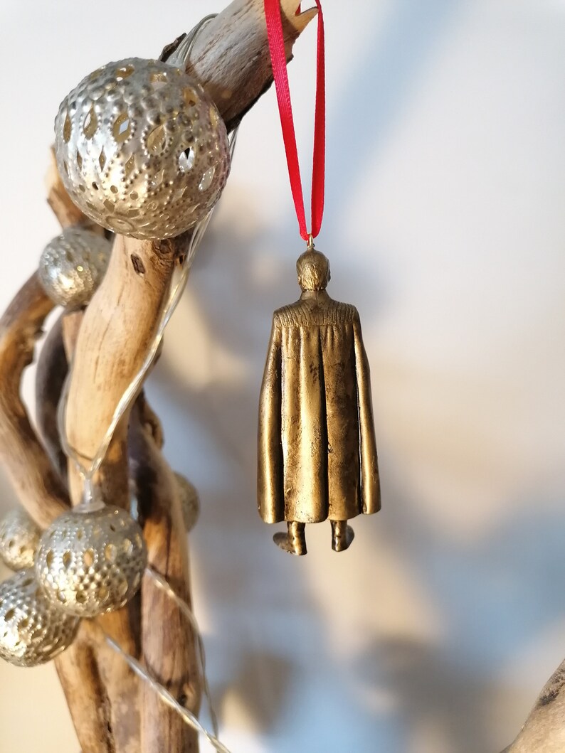 May include: A gold-colored ornament of a person wearing a cape, hanging from a red ribbon.
