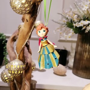 Pu&ograve; includere: Una piccola figurina di plastica di una ragazza con i capelli rossi, che indossa un vestito blu e oro con un mantello rosso. La figurina &egrave; appesa a uno spago verde.