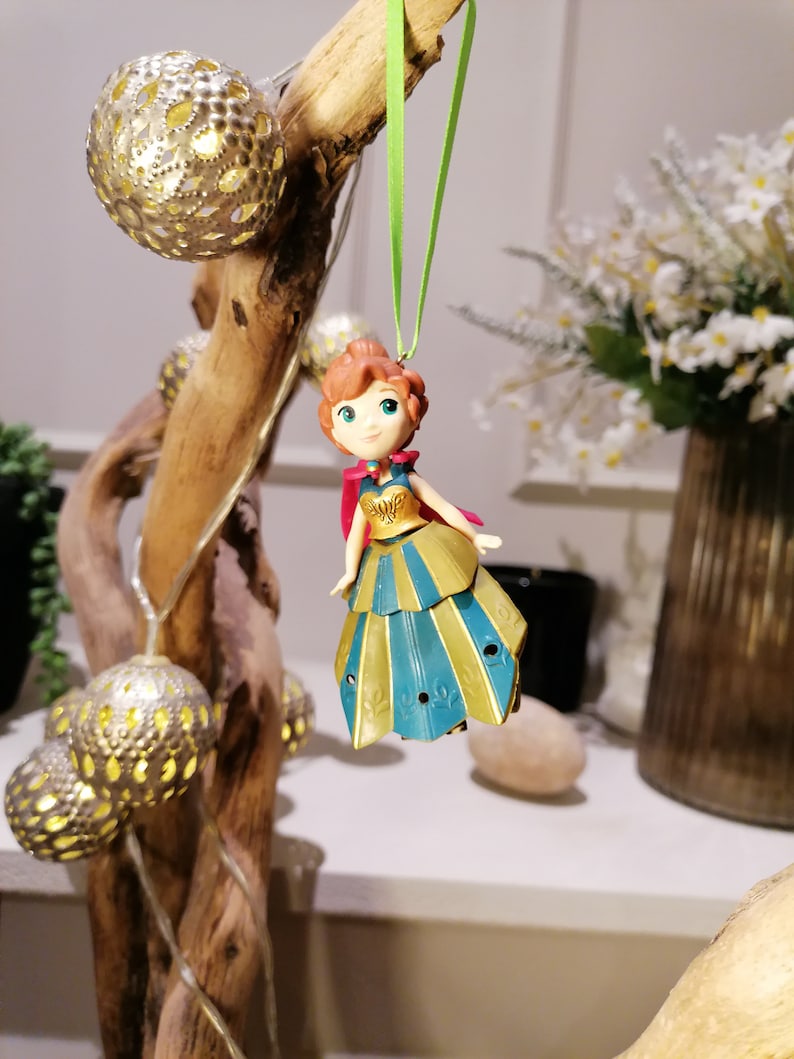 Pu&ograve; includere: Una piccola figurina di plastica di una ragazza con i capelli rossi, che indossa un vestito blu e oro con un mantello rosso. La figurina &egrave; appesa a uno spago verde.