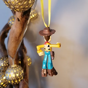 May include: A Woody action figure Christmas ornament. The toy cowboy has a brown hat, yellow shirt, blue pants, and brown boots. It is hanging from a yellow ribbon. The background includes a wooden branch and string lights.
