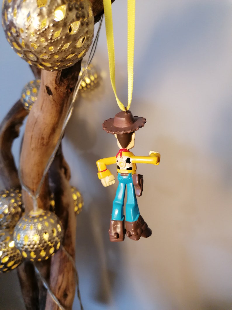 May include: A Woody action figure Christmas ornament. The toy cowboy has a brown hat, yellow shirt, blue pants, and brown boots. It is hanging from a yellow ribbon. The background includes a wooden branch and string lights.