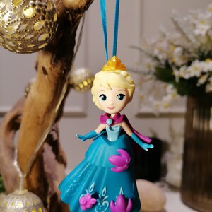 K&ouml;nnte beinhalten: Ein blauer und lilafarbener Plastik-Anh&auml;nger einer Prinzessin in einem blauen Kleid und einer goldenen Krone. Der Anh&auml;nger h&auml;ngt an einem blauen Band.
