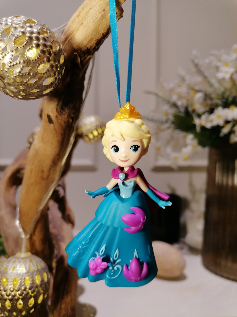 K&ouml;nnte beinhalten: Ein blauer und lilafarbener Plastik-Anh&auml;nger einer Prinzessin in einem blauen Kleid und einer goldenen Krone. Der Anh&auml;nger h&auml;ngt an einem blauen Band.
