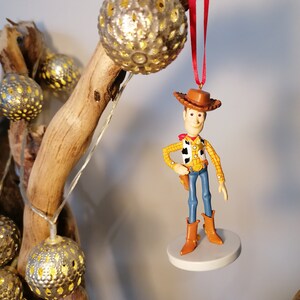 Op de afbeelding: Een rode draad hangt aan een houten tak met een zilveren ornament. Een plastic figuurtje van Woody uit Toy Story hangt aan de draad. Woody draagt zijn kenmerkende cowboyhoed, vest en jeans.