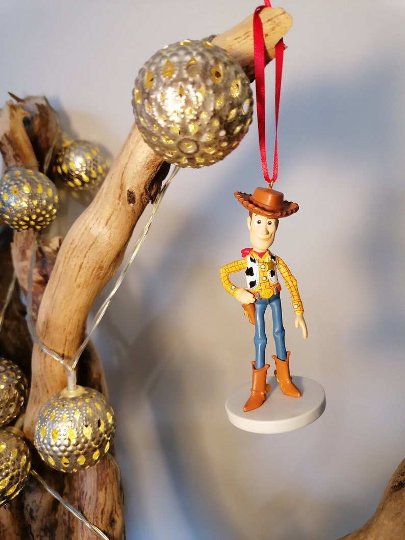 Op de afbeelding: Een rode draad hangt aan een houten tak met een zilveren ornament. Een plastic figuurtje van Woody uit Toy Story hangt aan de draad. Woody draagt zijn kenmerkende cowboyhoed, vest en jeans.