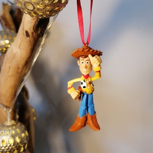 Pu&ograve; includere: Uno spago rosso pende da un ramo con una statuetta di plastica di Woody di Toy Story. Woody indossa il suo cappello da cowboy, il gilet e i jeans caratteristici. Si sta tenendo la mano destra alla testa e sorride.