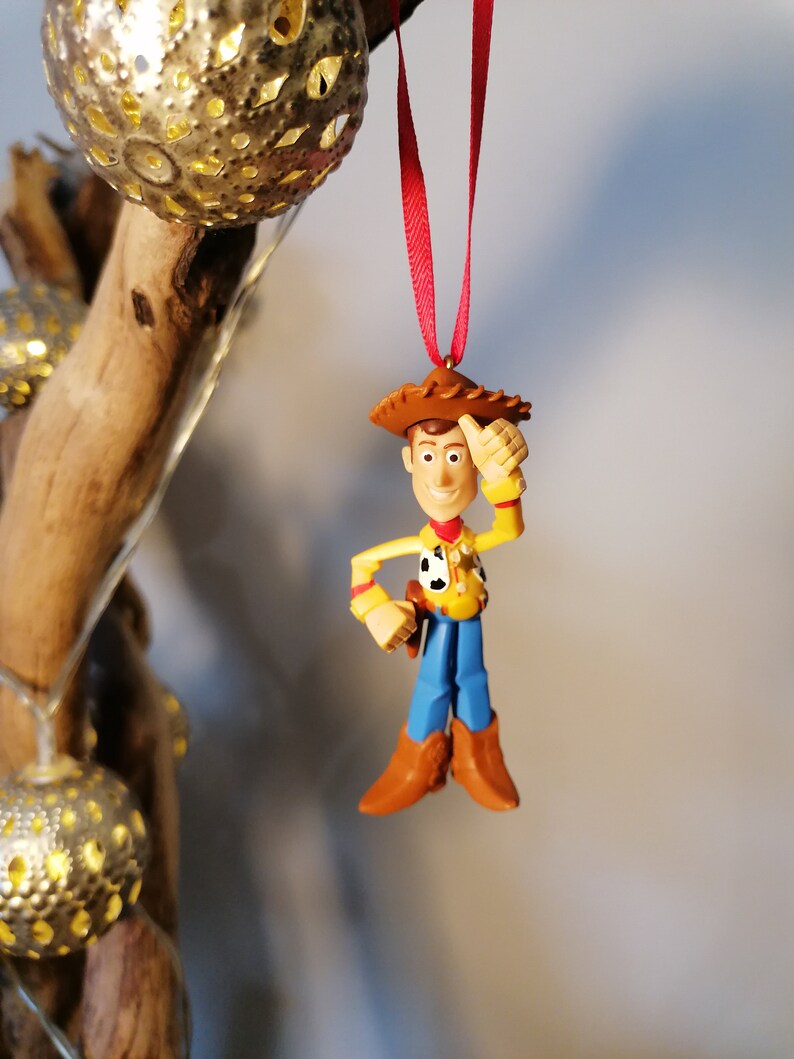 Pu&ograve; includere: Uno spago rosso pende da un ramo con una statuetta di plastica di Woody di Toy Story. Woody indossa il suo cappello da cowboy, il gilet e i jeans caratteristici. Si sta tenendo la mano destra alla testa e sorride.