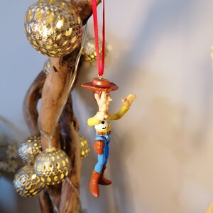 May include: A red string hangs from a branch with a plastic toy figurine of Woody from Toy Story. Woody is wearing his signature cowboy hat, vest, and jeans. He is holding his right arm out and smiling.