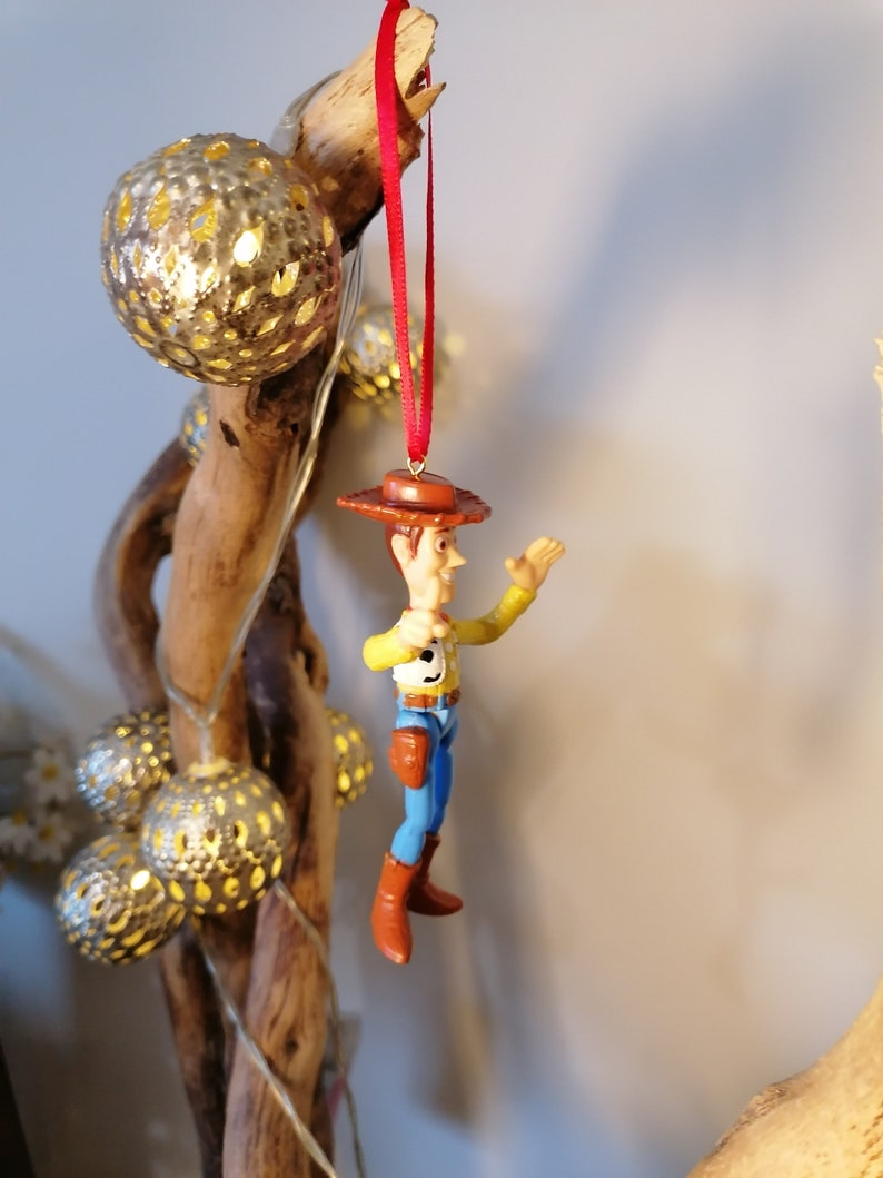 May include: A red string hangs from a branch with a plastic toy figurine of Woody from Toy Story. Woody is wearing his signature cowboy hat, vest, and jeans. He is holding his right arm out and smiling.