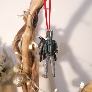May include: A green and gray Star Wars Boba Fett action figure ornament hanging from a red ribbon on a branch with white lights.