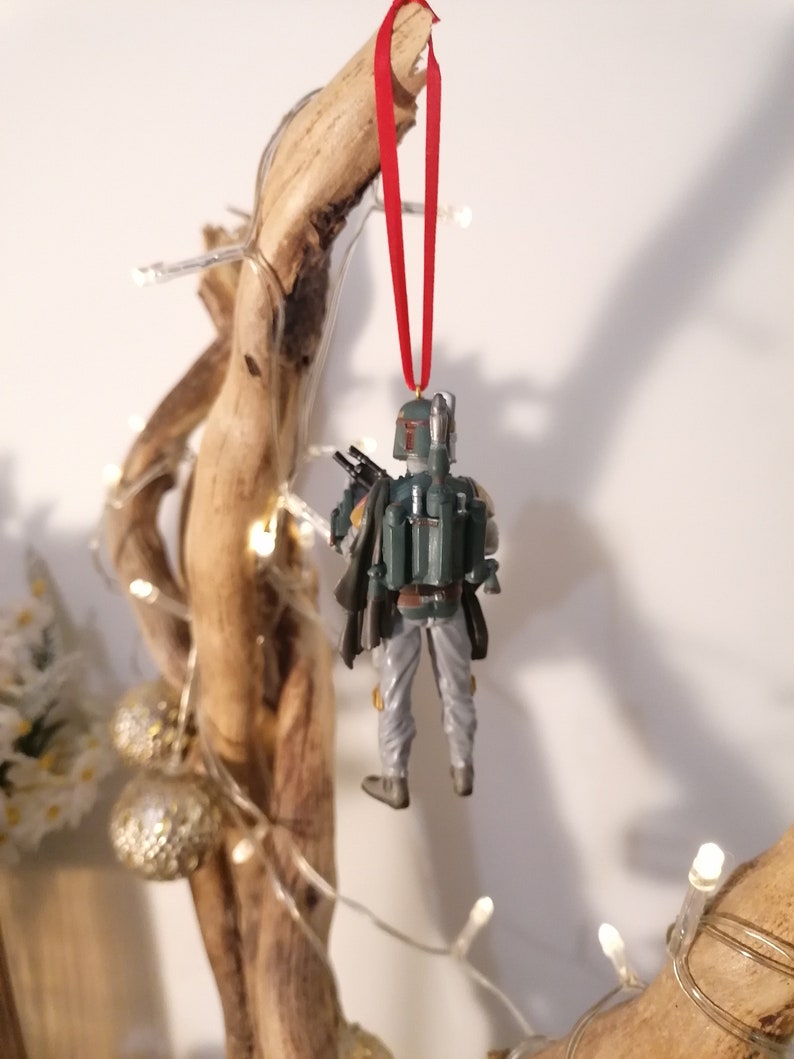 May include: A green and gray Star Wars Boba Fett action figure ornament hanging from a red ribbon on a branch with white lights.