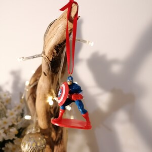 May include: A red, white, and blue Captain America Christmas ornament. The ornament features Captain America standing on a red surfboard, holding his shield in his left hand. The ornament is hanging from a red ribbon.