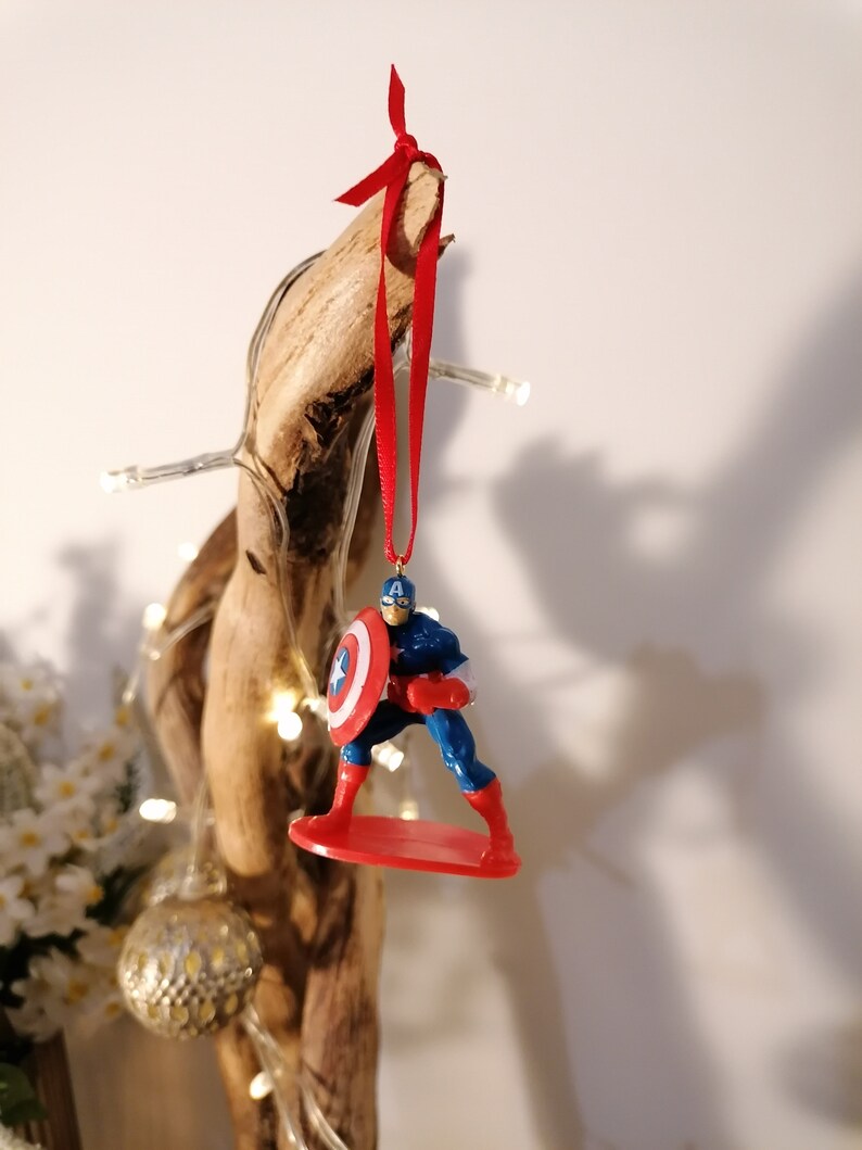 May include: A red, white, and blue Captain America Christmas ornament. The ornament features Captain America standing on a red surfboard, holding his shield in his left hand. The ornament is hanging from a red ribbon.