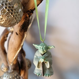 May include: A green Yoda ornament hanging from a light green ribbon. The ornament is made of plastic and features Yoda holding a staff.